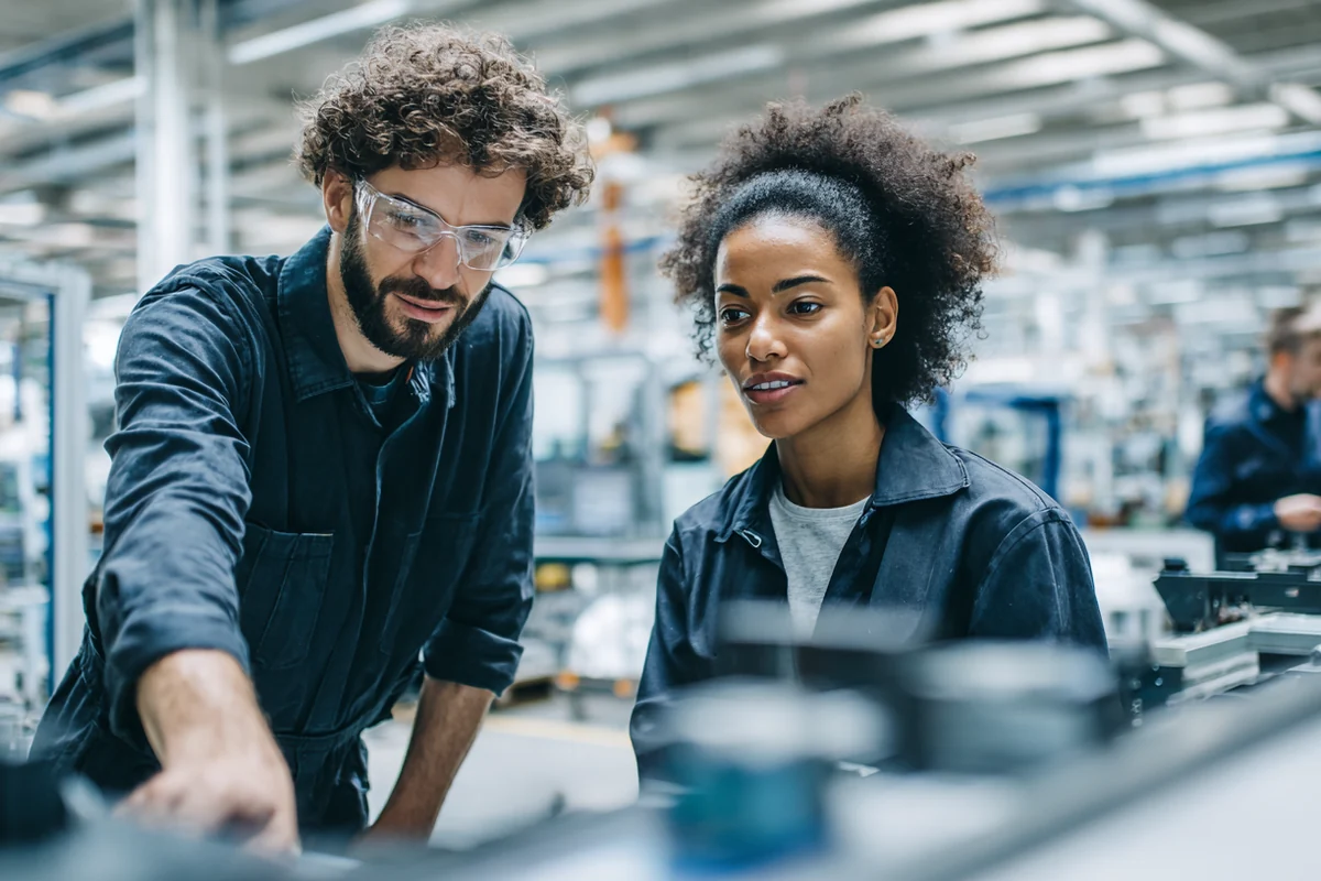 Dos técnicos revisando una máquina en una planta de manufactura, con la línea de producción al fondo